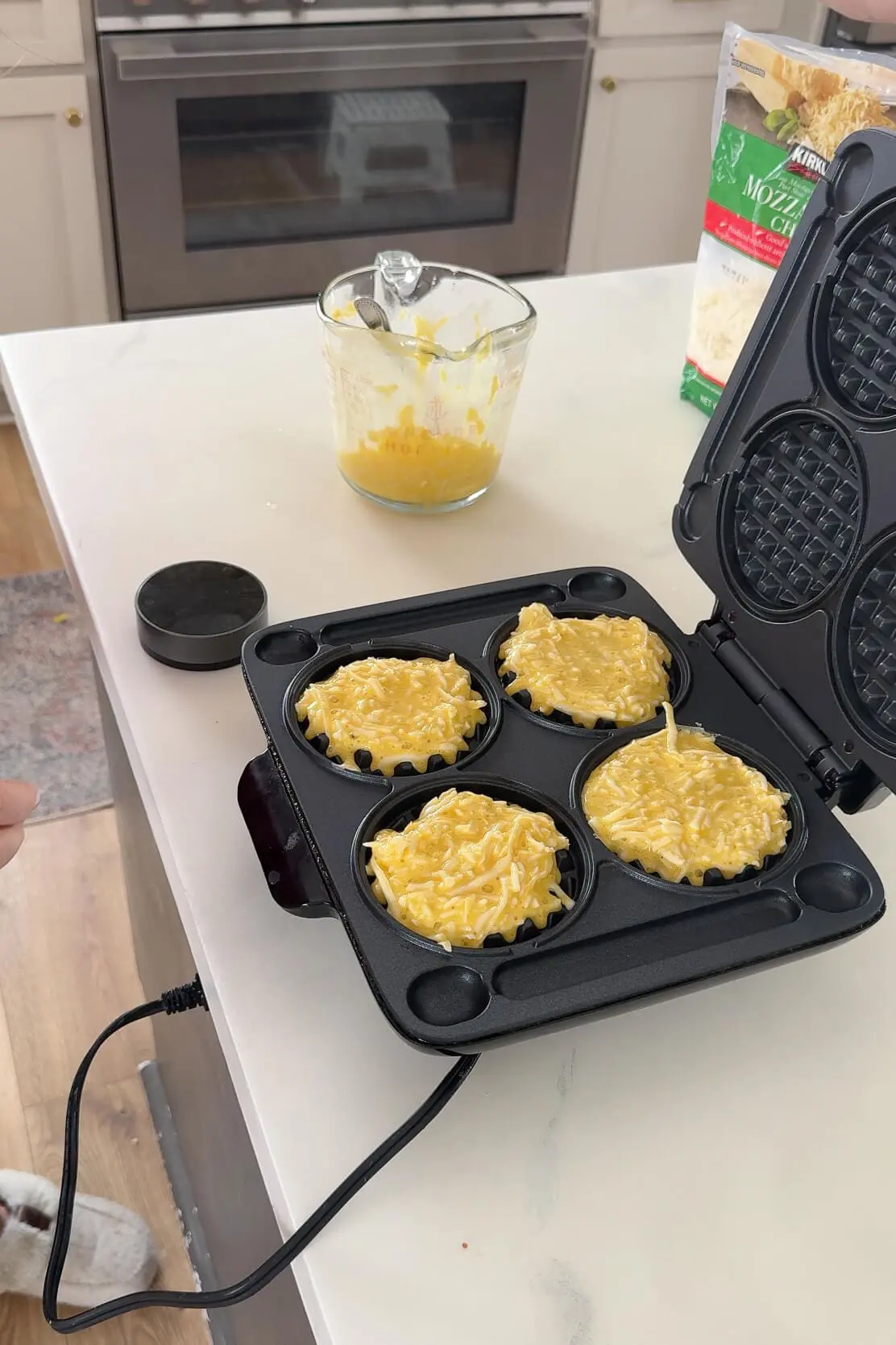 A close-up of an open waffle maker on a white kitchen countertop, filled with four uncooked chaffles being made as a carnivore bread alternative. A measuring cup with leftover batter and a bag of shredded mozzarella cheese sit nearby, indicating the chaffle-making process.